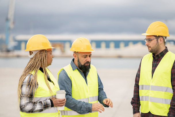 Diversi team di costruzione impegnati in una discussione in un porto, con una donna afroamericana, uomo del Medio Oriente e uomo caucasico in equipaggiamento di sicurezza. - Foto, immagini