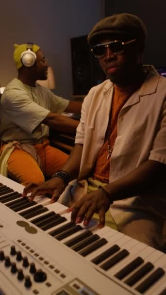 Vertical médio do músico afro-americano de boné, óculos de sol tocando teclado no piano elétrico no estúdio de gravação, engenheiro de áudio sorridente ouvindo e operando placa de mistura - Filmagem, Vídeo