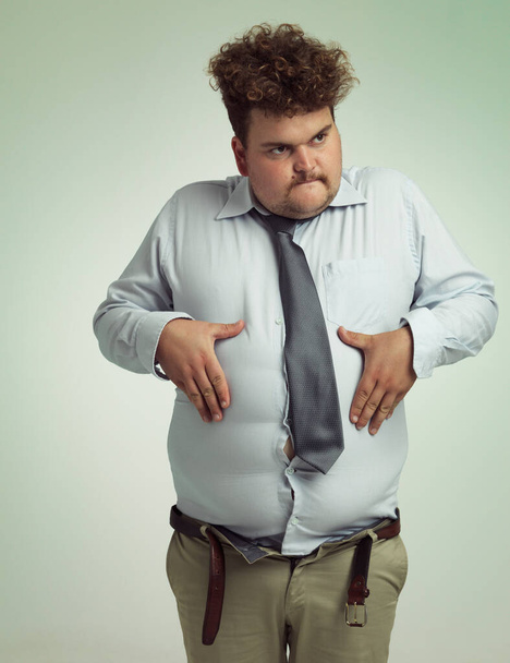 Człowiek, żołądek i plus rozmiar z brzucha z niezdrowej diety lub przyrost masy ciała na tle studio. Młody mężczyzna lub duży facet trzymający brzuch za zaburzenia jedzenia lub świadomość ciała na przestrzeni makiety. - Zdjęcie, obraz