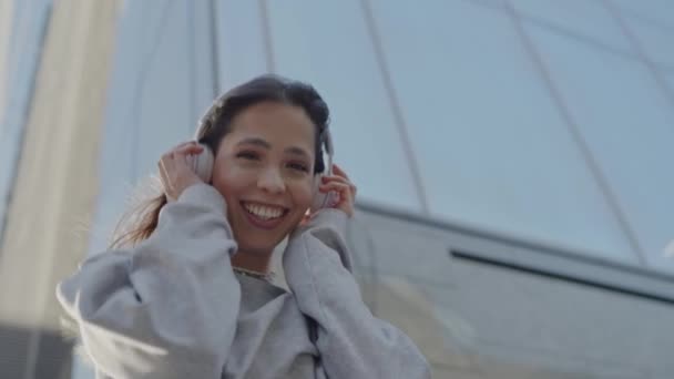 Μια χαρούμενη νεαρή γυναίκα ακούει μουσική με ακουστικά, χαμογελώντας χαρούμενα σε μια φωτεινή αστική μέρα. Νεαρή γυναίκα απολαμβάνει τη μουσική στο αστικό περιβάλλον, ηλιόλουστη μέρα - Πλάνα, βίντεο