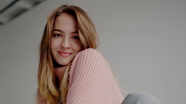 A portrait of a young, joyful woman in a cozy pink sweater, smiling warmly wrapped in her arms on a neutral background. Perfect for themes of comfort and happiness. Young Woman in Pink Sweater Smiling - Footage, Video