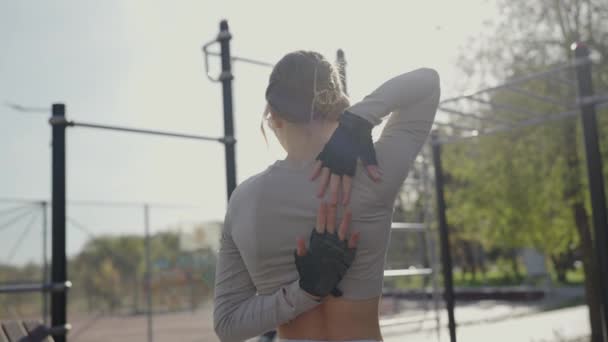 Pohled zezadu na mladou ženu, jak si natahuje ruce, připravuje se nebo dokončuje cvičení v parku. Sluneční světlo posiluje klidné prostředí. Mladá žena roztahování po venkovní cvičení v ranním světle - Záběry, video