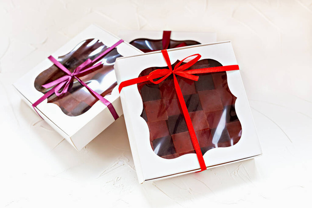 Tři sofistikované bílé dárkové krabice s průsvitnými okny s pestrobarevnými marmeládami, zdobené stylovými červenými a fialovými stuhami, ideální pro zvláštní příležitosti. Ručně vyráběné cukrovinky - Fotografie, Obrázek