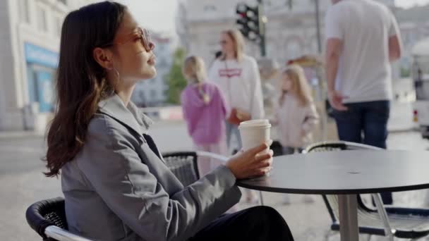 Eine junge Frau in professioneller Kleidung genießt einen friedlichen Moment in einem Café im Freien und hält eine Kaffeetasse in der Hand, während Menschen in der Stadt vorbeigehen. Junge Berufstätige entspannen sich im Outdoor-Café in der Stadt - Filmmaterial, Video
