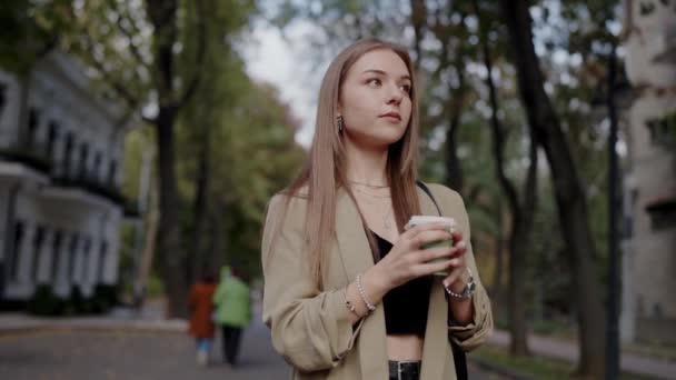Portré egy fiatal nőről, aki egy vibráló őszi parkban sétál, miközben egy csésze kávét tart a kezében. Figyelmes kifejezés, alkalmi bézs dzsekibe öltözve. Fiatal nő kezében kávé séta egy városi parkban - Felvétel, videó