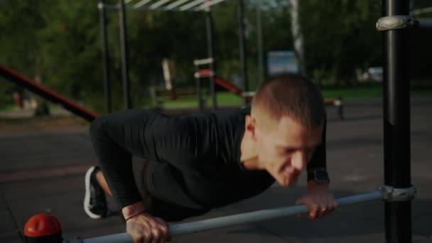Atletický mladý muž předvádí kliky na baru ve fitness parku, demonstruje sílu a vytrvalost během venkovního cvičení. Mladý muž cvičí venkovní Push-Ups ve fitness parku - Záběry, video