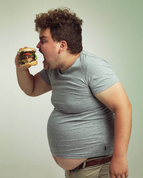 Ожирение, увеличение веса и фаст-фуд для человека в студии с гамбургером для нездорового питания, а также размер и жадность. Избыточный вес человека с едой для рта и жира желудка для голода, комедии и укуса в помещении. - Фото, изображение