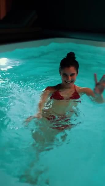 Una joven en traje de baño nada en una piscina cubierta con iluminación. Concepto de vacaciones del hotel. Vídeo vertical - Imágenes, Vídeo