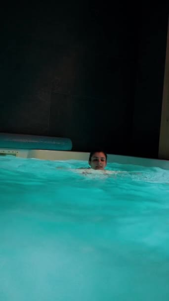 Молодая женщина в купальнике плавает в закрытом бассейне с освещением. Концепция отдыха в отеле. Вертикальное видео - Кадры, видео