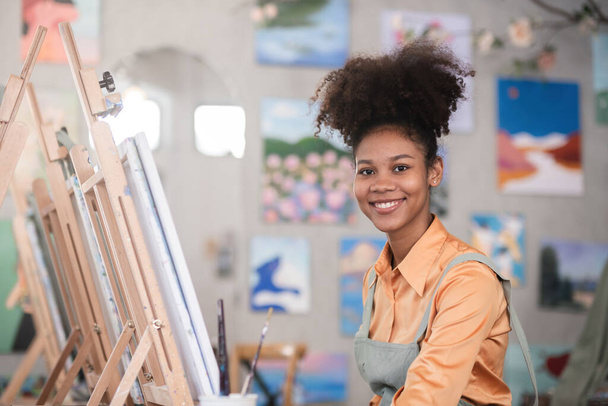 Μια νεαρή μαύρη καλλιτέχνης ζωγραφίζει ακρυλικά χρώματα σε καμβά με αποφασιστικότητα στο εργαστήριό της ζωγραφικής. - Φωτογραφία, εικόνα
