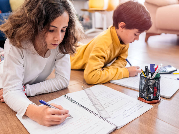 Duas crianças escrevendo em cadernos, cercadas por lápis e uma calculadora, focada em seus trabalhos de casa. - Foto, Imagem