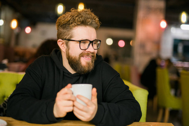 Γενειοφόρος σγουρά άνθρωπος χαμογελά αυτοπεποίθηση πίνοντας καφέ στο εστιατόριο ή την καφετέρια. Χιλιετηρίδα και Γενιά Y. - Φωτογραφία, εικόνα