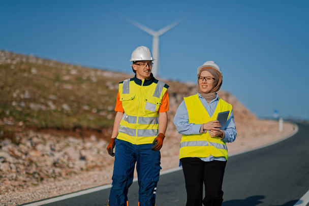 Una ingeniera con una tableta y su colega inspeccionan turbinas eólicas en un campo, demostrando trabajo en equipo y dedicación a proyectos de energía renovable.  - Foto, imagen