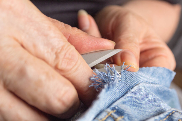 Bliżej dłoni kobiety używającej nożyczek do postrzępienia kawałka jeansowej tkaniny. Koncepcja transformacji odzieży. Krawiectwo. - Zdjęcie, obraz