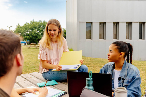многорасовая группа студентов с акцентом на молодую белую девушку, обучающуюся вместе, сидящую за столом за пределами кампуса колледжа - Фото, изображение