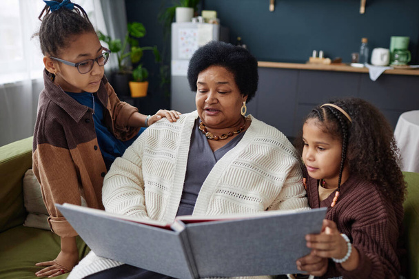 Retrato de una mujer negra mayor con dos hijos mirando el álbum de fotos juntos y enseñando historia familiar - Foto, Imagen