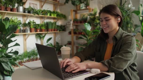 Μέτριο πλάνο του Καυκάσου Γυναίκα επιχειρηματίας της κηπουρικής επιχείρηση που εργάζονται σε φορητό υπολογιστή χρησιμοποιώντας ακουστικά και μιλώντας με τους πελάτες, πράσινο γλάστρες φυτά στα ράφια στο παρασκήνιο - Πλάνα, βίντεο
