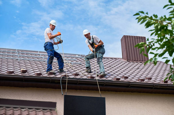 Hommes travailleurs installant le système de panneaux solaires sur le toit de la maison. Electriciens dans les casques soulevant le module solaire photovoltaïque à l'aide de cordes à l'extérieur. Concept d'énergies alternatives et renouvelables. - Photo, image