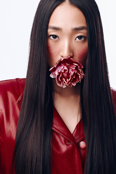Atractiva joven con el pelo largo y negro y chaqueta roja posando con flor en la boca - Foto, Imagen