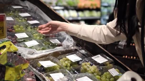 In un vivace supermercato, una donna seleziona l'uva, ispezionando ogni confezione per l'uva più fresca. Controlla attentamente la qualità delle uve, assicurandosi che le uve che sceglie siano grasse e succose. - Filmati, video
