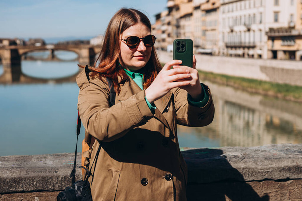 Aantrekkelijke jonge vrouwelijke toerist verkent nieuwe stad. Roodharig meisje met een smartphone op de beroemde Oude brug in Florence. Reizen. gelukkig meisje wandelen in de stad en maakt selfie - Foto, afbeelding