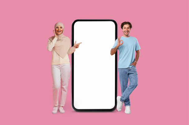 Una mujer en hijab y el hombre apuntan a la pantalla del teléfono inteligente en blanco sobre un fondo rosa, espacio de copia de maqueta, aplicación móvil - Foto, imagen