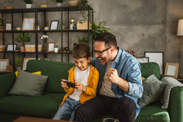 Vader en zoon spelen videospelletjes op een mobiele telefoon en genieten thuis - Foto, afbeelding
