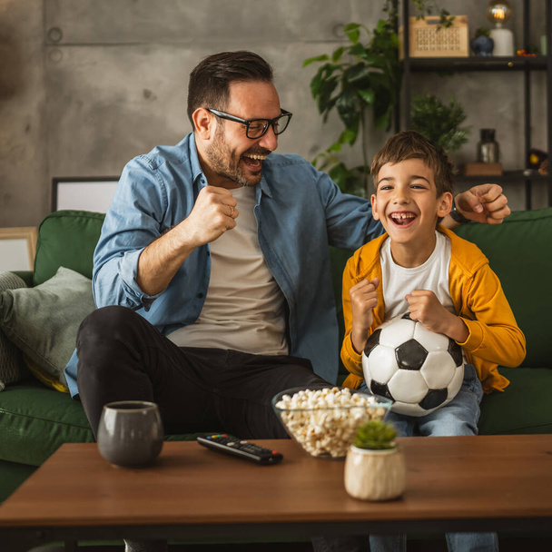 Πατέρας και γιος καυκάσιος παρακολουθούν ποδοσφαιρικό αγώνα και ζητωκραυγάζουν στο σπίτι θορυβώδεις, ενθουσιασμένοι, ευτυχισμένοι - Φωτογραφία, εικόνα