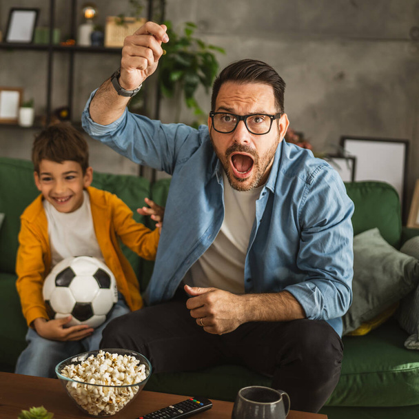 Ojciec i syn kaukaski oglądać mecz piłki nożnej i dopingować w domu hałaśliwy, podekscytowany, szczęśliwy - Zdjęcie, obraz