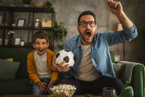 Батько і син кавказький дивитися футбольний матч і підбадьорювати вдома галасливий, збуджений, щасливий - Фото, зображення