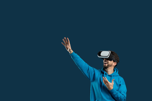 Ein Mann mit blauem Kapuzenpullover taucht in eine Virtual-Reality-Erfahrung ein, während er ein Headset trägt. Er interagiert mit einer digitalen Umgebung, während er sich mit der virtuellen Welt auseinandersetzt - Foto, Bild