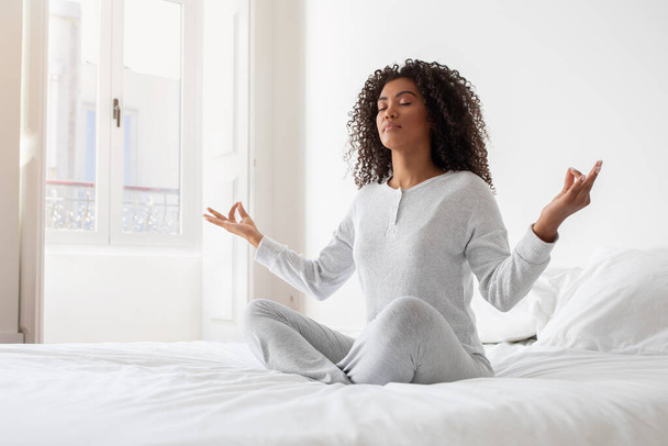 Een serene jonge Spaanse vrouw zit gekruist op een wit bed, met haar ogen dicht en handen in een meditatieve houding. Soft morning light filtert door het raam - Foto, afbeelding