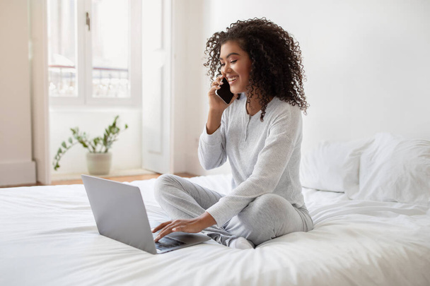 Una giovane donna ispanica è seduta su un letto bianco mentre si impegna in una conversazione telefonica e lavora sul suo computer portatile. La stanza è ben illuminata con luce naturale - Foto, immagini