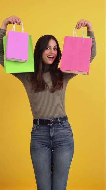 Vertical. Happy young adult caucasian woman enjoying and carrying shopping bags over yellow or orange background. Fashion female smiling showing her sale purchase. Customer lady advertsing retail day - Footage, Video