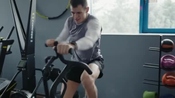 Εστιασμένος νεαρός άνδρας δίνει τα πάντα, ενώ ποδηλατεί σε ένα σύγχρονο σταθερό ποδήλατο σε ένα καλά εξοπλισμένο γυμναστήριο, που περιβάλλεται από εξοπλισμό γυμναστικής. Νεαρός άνθρωπος εκπαίδευση έντονα σε ένα σταθερό ποδήλατο στο γυμναστήριο - Πλάνα, βίντεο