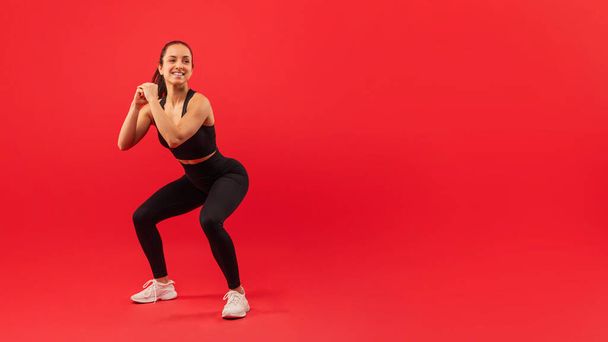 A joyful woman wearing sportswear, including a black tank top and leggings, performs a squat exercise with her hands together, red background, copy space - Photo, Image