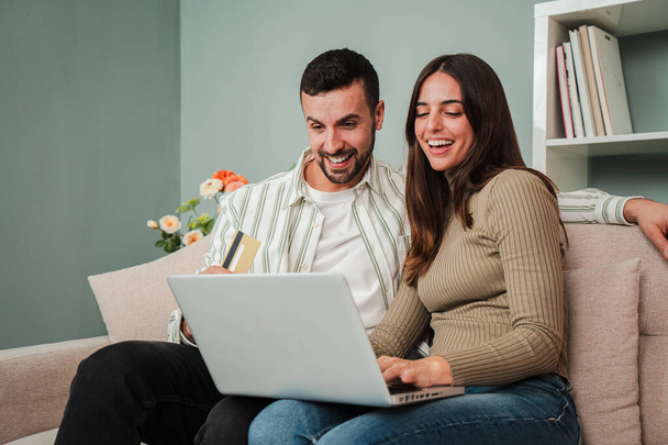 Boldog fiatal kaukázusi házaspár böngésznek az interneten egy laptop segítségével, hogy keressen egy eladó otthon. Izgatott férj és feleség mosolyog, és keres egy új lakást ül egy kanapén a nappaliban. Magas - Fotó, kép