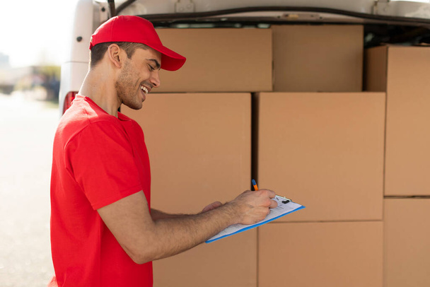 Professional young delivery man in uniform checking delivery list before handed to his customer, standing next to van full of parcel boxes - Photo, Image