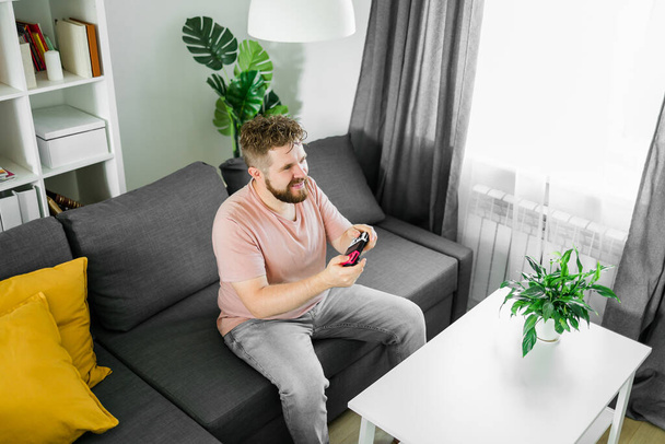 Молодий чоловік проводить час вдома, сидячи на дивані в квартирі і граючи в аркадні відеоігри на консолі. Чоловік за допомогою контролера для відтворення симулятора дрейфу вуличних гонок - Фото, зображення
