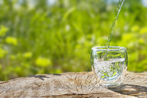 Woda przelewana jest do szklanki na tle zieleni. Szklanka wody w upalny letni dzień z przestrzenią do skopiowania. - Zdjęcie, obraz