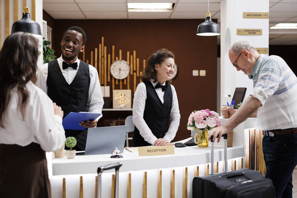 Retired senior couple arrive at hotel, assisted by smiling multiethnic staff at front desk. Check-in, paperwork, luggage. Luxury hotel lobby bustling with tourists. Excited for vacation. - Photo, Image