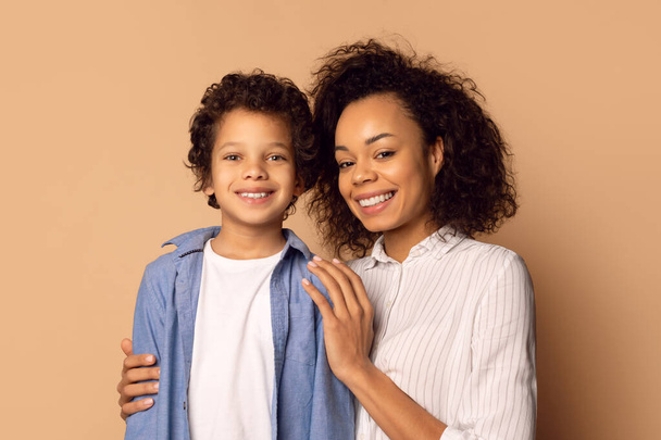 Femme afro-américaine et un enfant sont debout ensemble, souriant et posant pour une photographie. La femme a son bras autour de l'enfant, fond beige - Photo, image