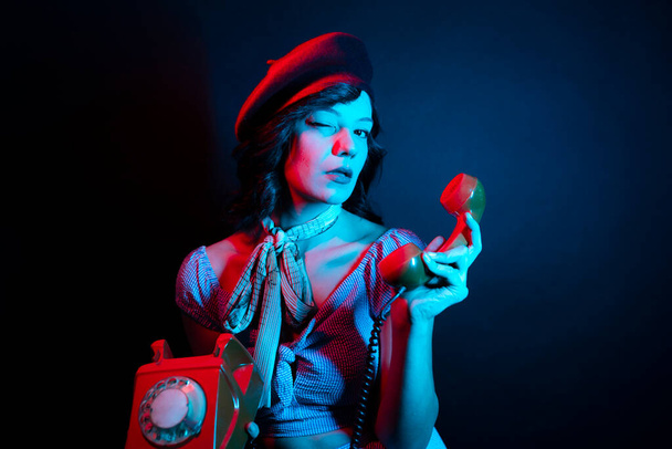 Νεαρή γυναίκα που μιλάει στο πορτοκαλί ρετρό τηλέφωνο. Υψηλή Μόδα μοντέλο γυναίκα σε πολύχρωμο φωτεινό νέον μπλε και μοβ φώτα ποζάρουν στο στούντιο. - Φωτογραφία, εικόνα