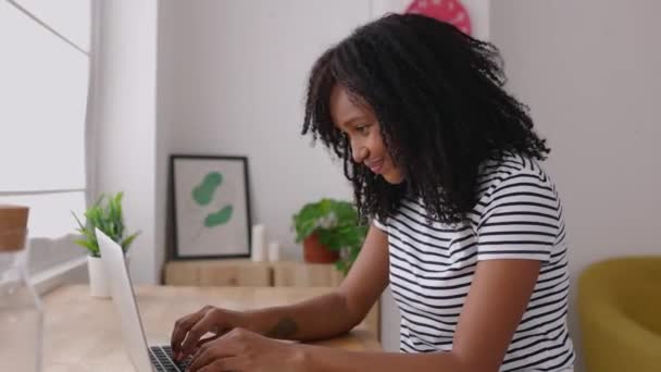 Young african american woman with curly hair working on her laptop computer while sitting on a desk at workplace. Happy millennial brazilian female studying online at home - Felvétel, videó