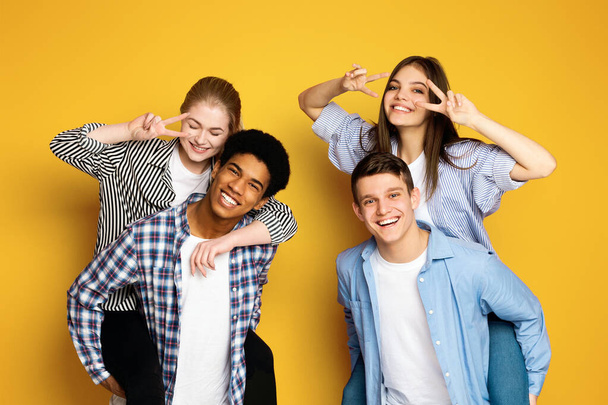 Un alegre grupo de cuatro jóvenes amigos posan juntos juguetonamente, con dos señales de paz cerca de sus ojos y todas sonrisas genuinas y alegres. - Foto, Imagen