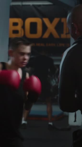 Függőleges lövés fókuszált fiatal harcos üt boxzsák edzés közben sötét box tornaterem. Atlétikai tinédzser boksz kesztyű gyakorlatok edző előtt harci torna. - Felvétel, videó
