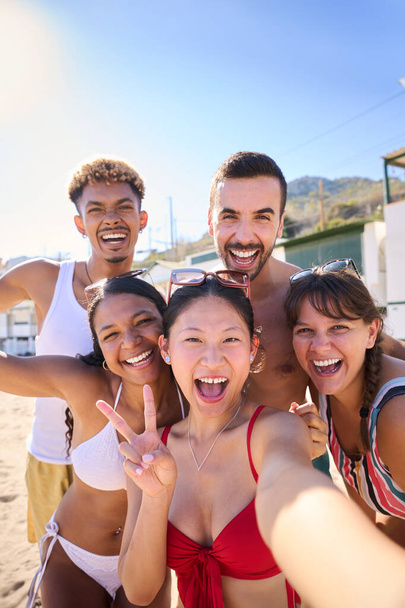 Група веселих багаторасових молодих друзів-генів, які беруть вертикальне селфі на пляжі, дивлячись на камеру, посміхаючись і розважаючись разом. Люди насолоджуються літнім відпочинком і подорожами у відпустку - Фото, зображення