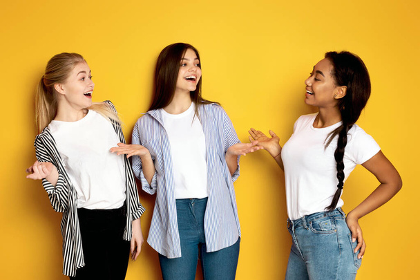 Tre ragazze multietniche stanno fianco a fianco con una di loro che fa gesti animati come nel bel mezzo di una storia coinvolgente, sfondo giallo dello studio - Foto, immagini