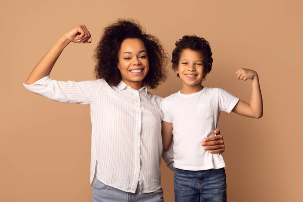 Uma mulher afro-americana sorridente e um menino estão flexionando seus músculos juntos, mostrando um vínculo de força e confiança. - Foto, Imagem