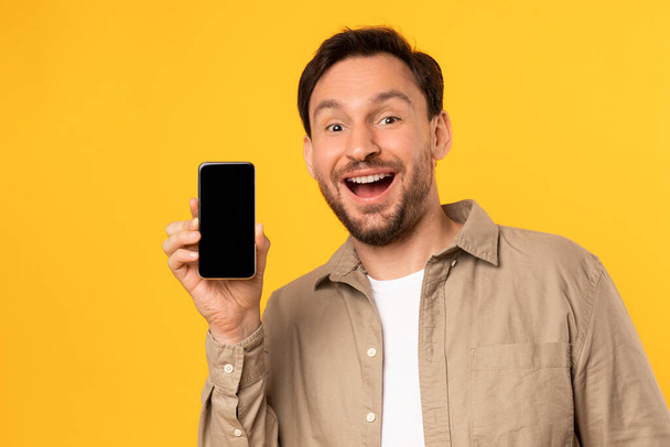 Ένας άντρας κρατάει ένα κινητό μπροστά στο πρόσωπό του, εστιάζοντας στην οθόνη. Φαίνεται να είναι απασχολημένος με τη συσκευή, δείχνει ωραία εφαρμογή ή online προσφορά - Φωτογραφία, εικόνα